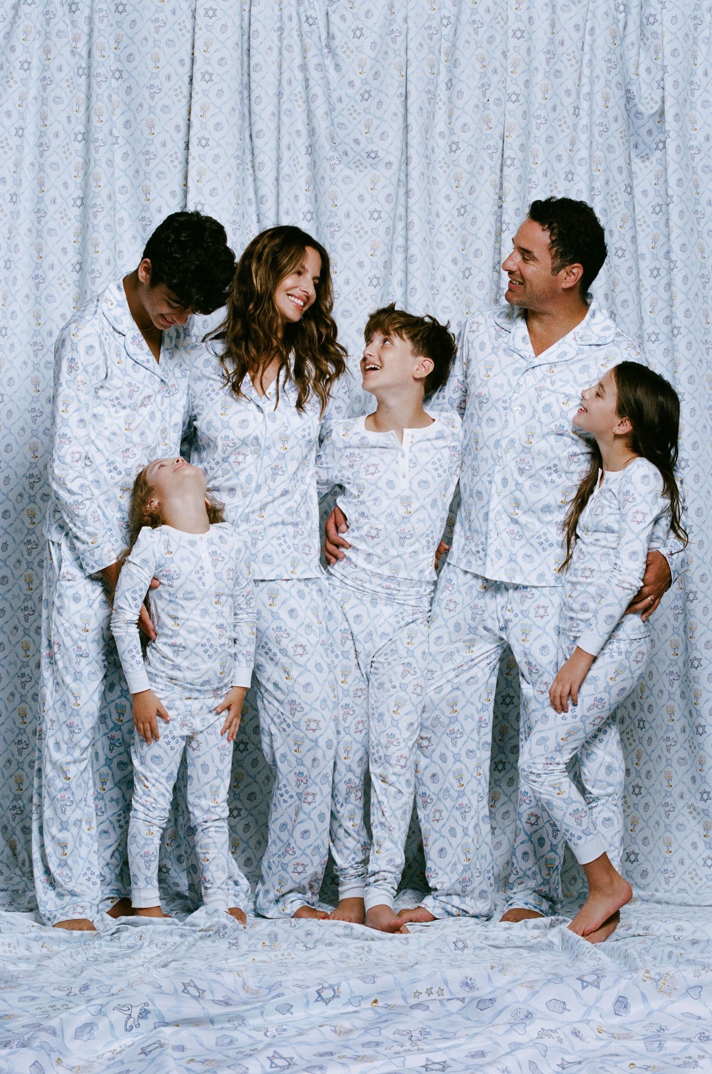 A man stands barefoot on a white background wearing Roller Rabbit's Mens Starry Nights Lattice Long Sleeve Polo Pajamas, featuring light blue and white patterns. He smiles with one hand in his pocket, highlighting the comfort of these matching PJs.