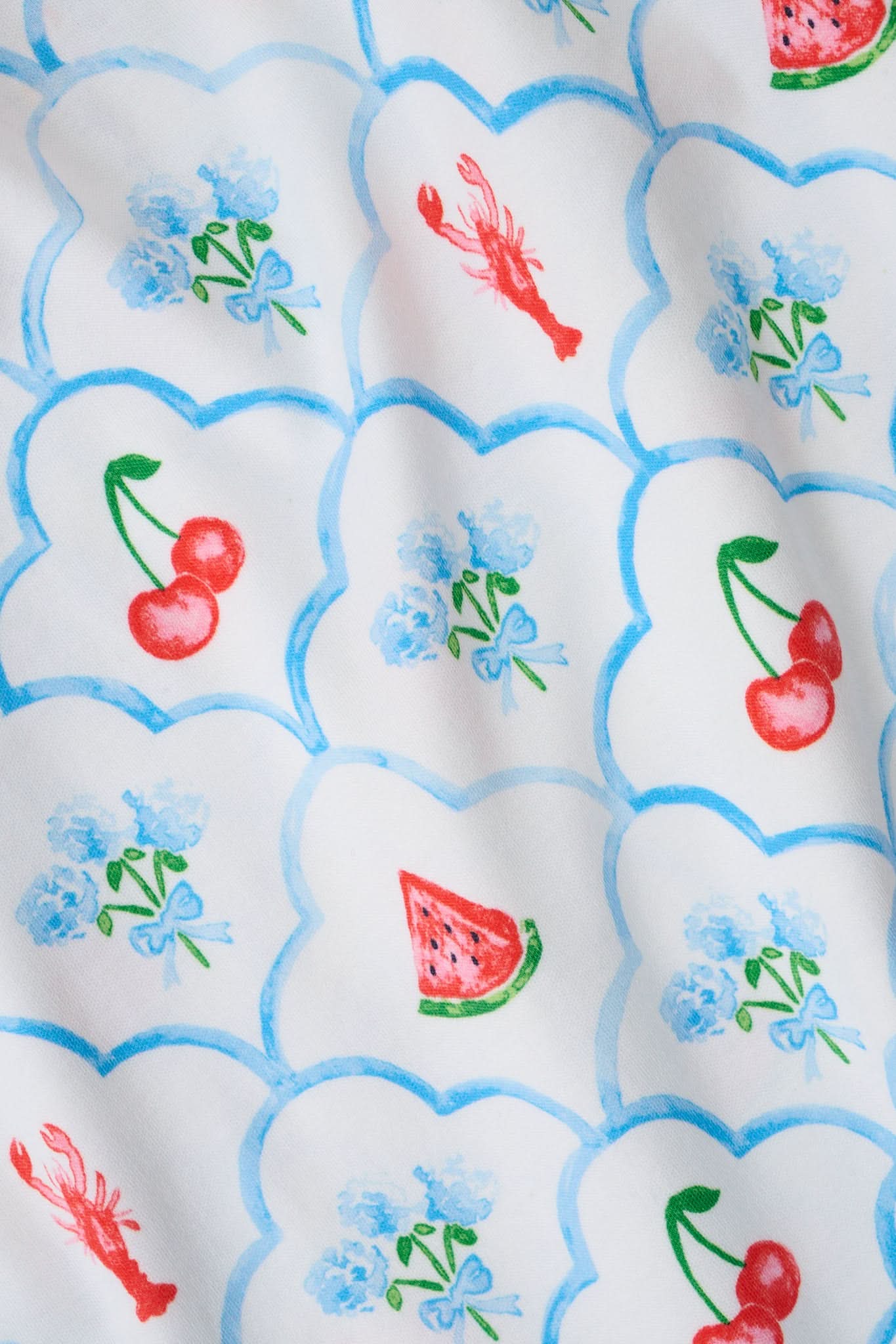 A woman with curly hair wears the Roller Rabbit 4th of Jemina Mischa Set, a Pima Cotton cami and shorts loungewear set with a white base, colorful fruit and flower prints, red trim, and drawstring shorts edged in red.