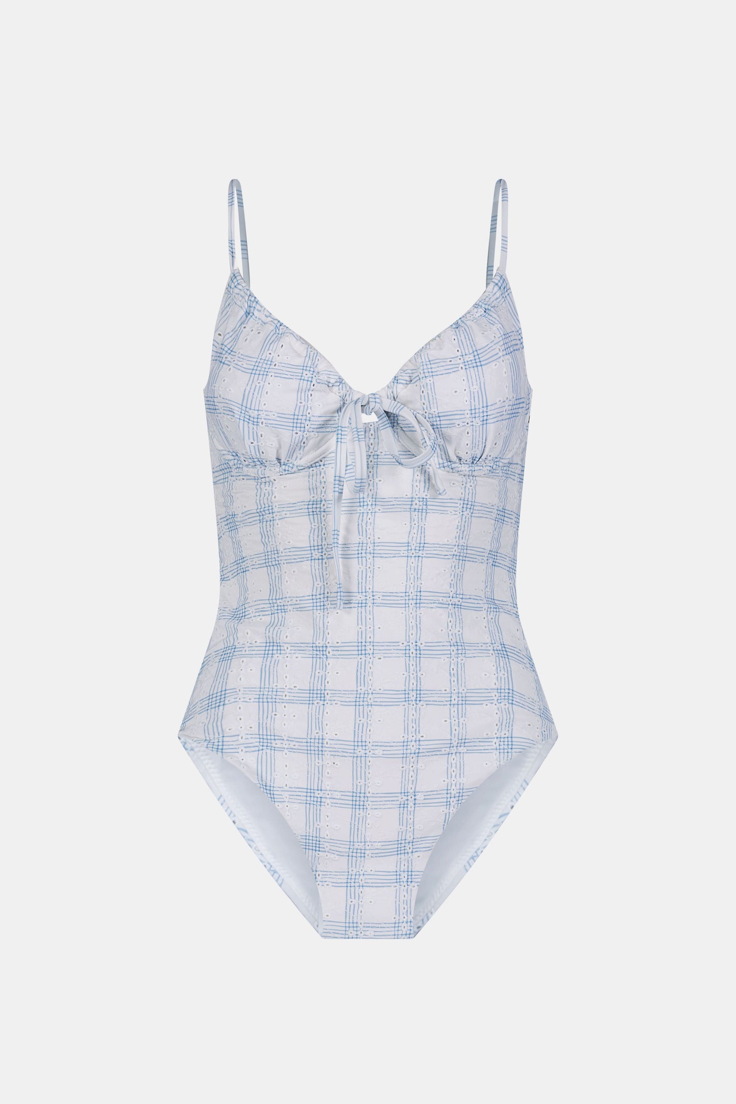 A woman wearing the Roller Rabbit Sun Check Eyelet Aurora One Piece, a light blue and white plaid swimsuit with adjustable straps, stands on a beach with waves and a blue sky in the background.
