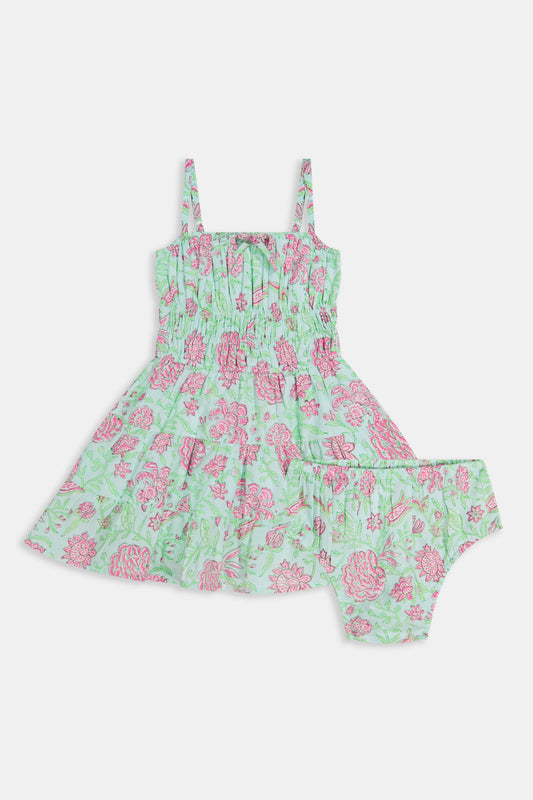 A young girl with long brown hair smiles, wearing the Roller Rabbit Kids Camellia Block Karla Dress, a sleeveless mint green cotton dress with pink floral patterns. She stands with one hand by her side and the other holding her dress.