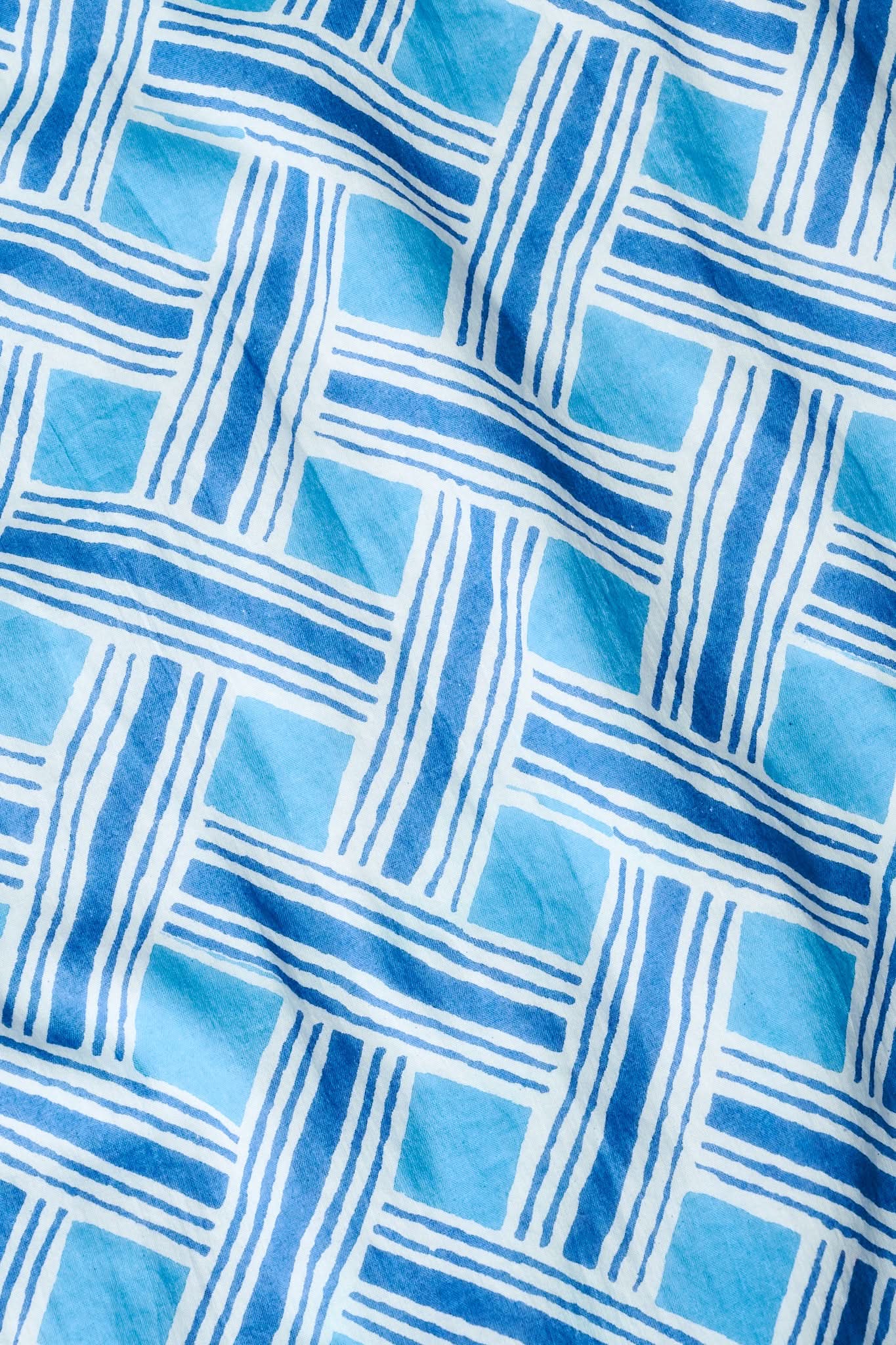 A close-up of the Liana Sabrina Top shows its cotton voile fabric with intersecting blue lines and rectangles on a light blue background, giving a woven, textured appearance—perfect for summer.