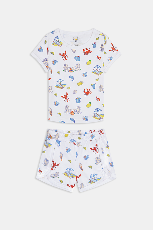 A young child smiles in a Roller Rabbit Kids Catch of the Day Playa Play Set—white t-shirt and shorts with colorful sea creature prints, paired with cream-colored shoes, against a plain white background.