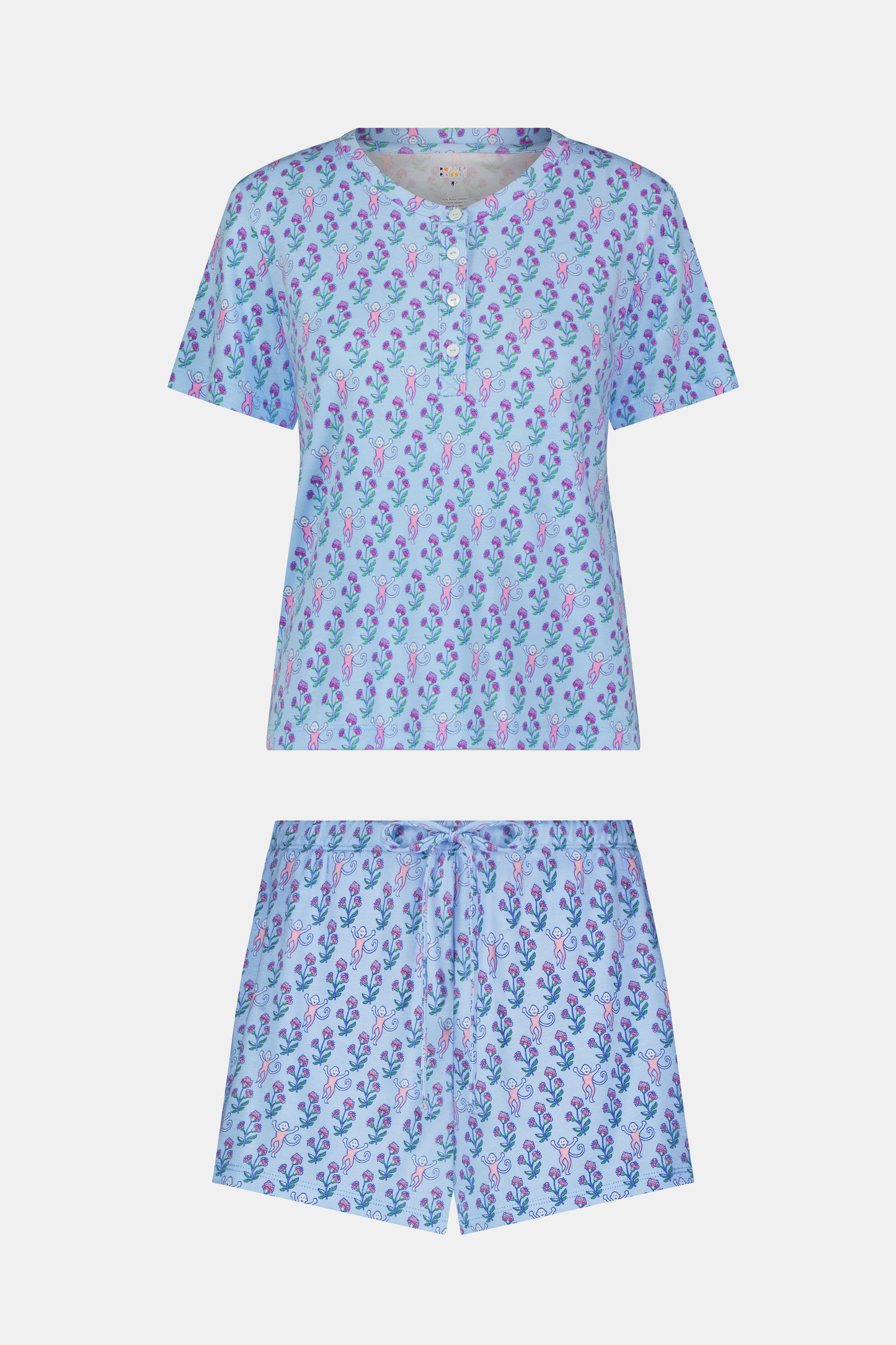 A woman with long curly hair wears the Rosa Ditsy Monkey Alexa Set by Roller Rabbit, a light blue Pima cotton pajama set featuring a ditsy monkey print. The set includes a henley-style top and matching shorts. She stands against a plain background.