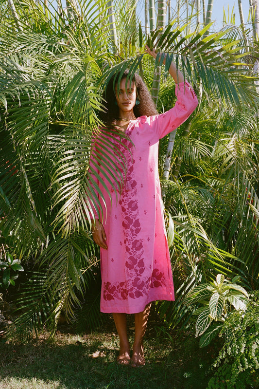A person stands barefoot among lush tropical plants, partly hidden by palm fronds, wearing the bright pink Cross Stitch Vine Shiv Caftan from Roller Rabbit as sunlight filters through the leaves.