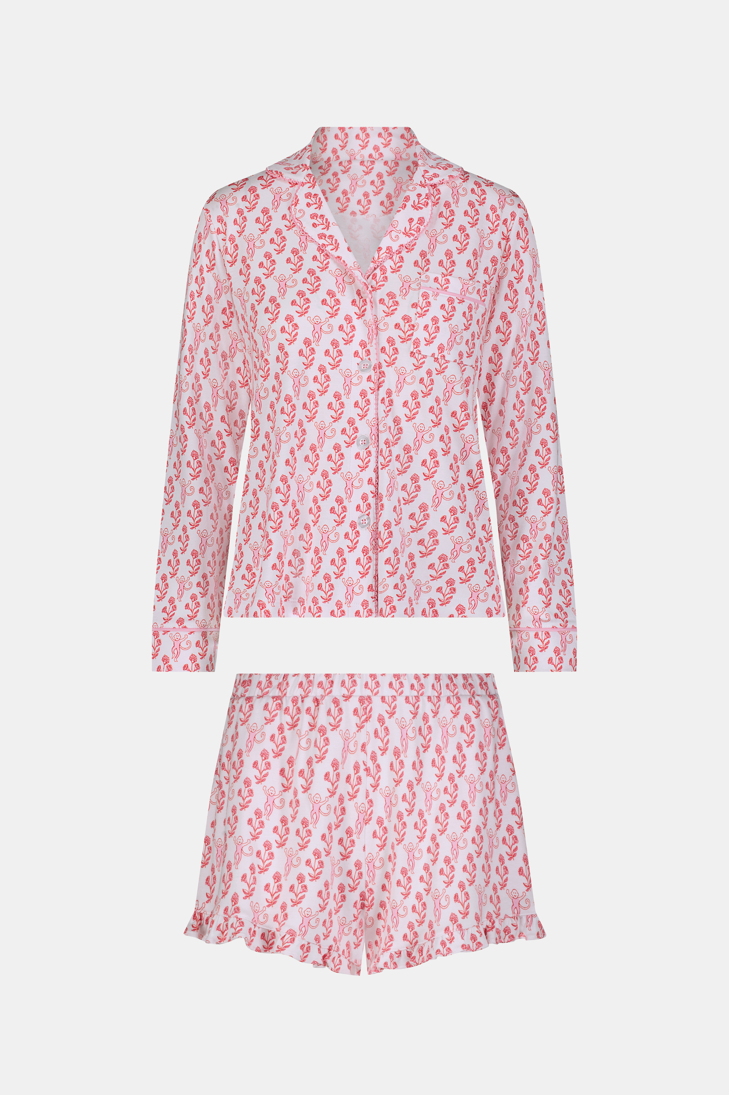 Wearing the Roller Rabbit Rosa Ditsy Monkey Hayden Pajamas—a pink and white set with a small anchor print, long-sleeve button-up top, and ruffled shorts—a person stands against a plain light background, smiling slightly.