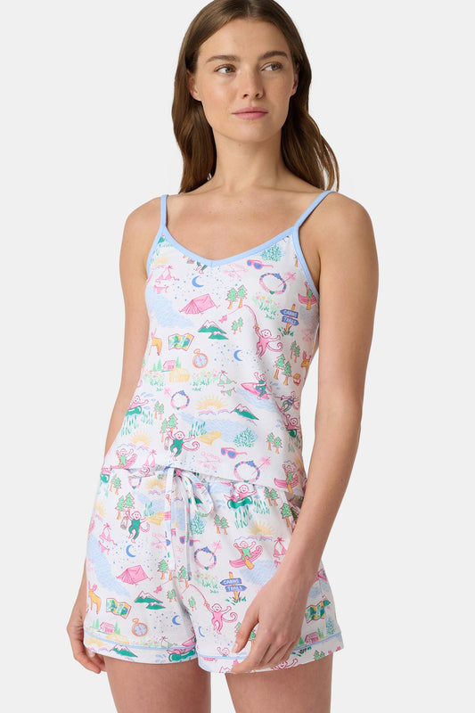 A woman with long brown hair wears the Roller Rabbit Camp RR Mischa Set, a light-hued Pima cotton pajama set with playful prints. The adjustable strap tank and shorts provide comfort as she stands turned slightly to the side, gazing off-camera.