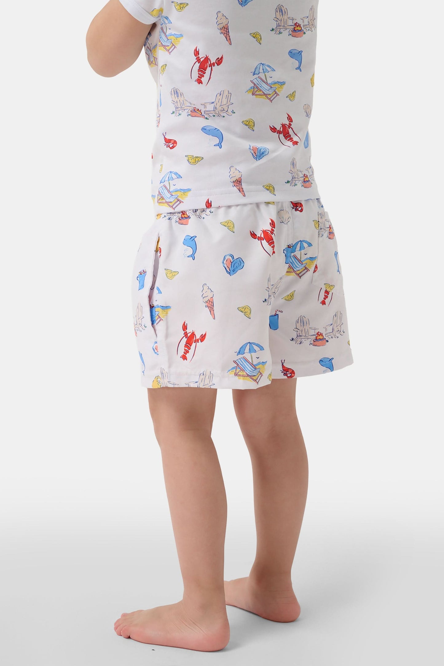 A young boy smiles barefoot in front of a cream-colored vehicle, wearing Roller Rabbit Boys Catch of the Day Shortie Swim Trunk with colorful prints, while another child sits partially visible beside him.
