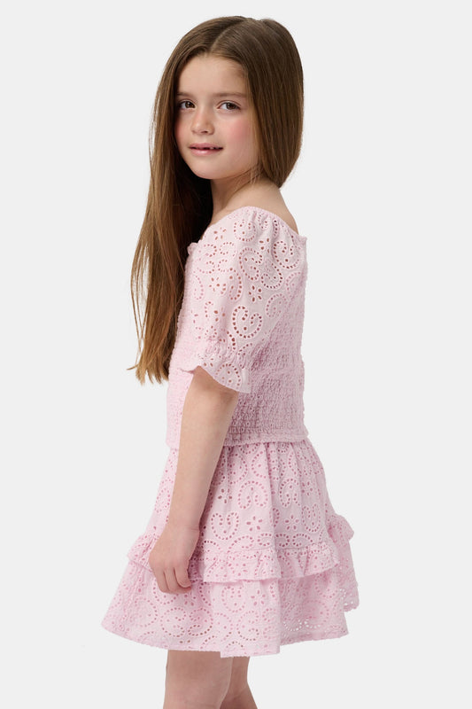 A young girl with long brown hair is wearing the Roller Rabbit Kids Clover Eyelet Elsie Top, a light pink, short-sleeved, smocked kids top, paired with a matching heart-patterned eyelet skirt. She smiles against a plain light background.