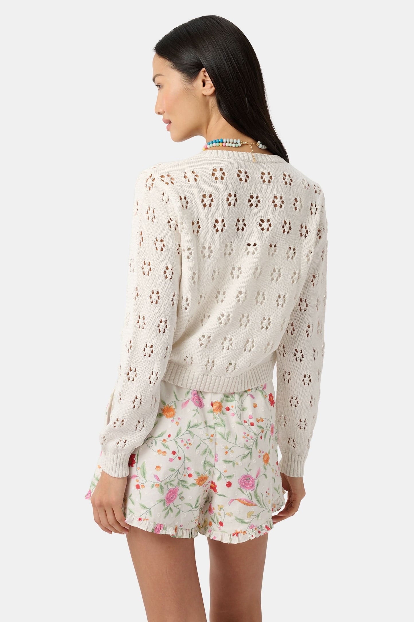 A woman with long dark hair smiles in the Roller Rabbit Floral Crochet Moxie Cardigan, featuring flower-like holes; she pairs it with light floral ruffled-hem shorts and rests one hand in her pocket.