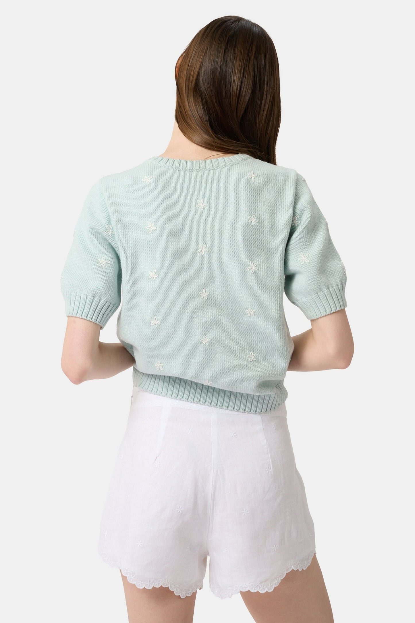 A person wears the Roller Rabbit Ditsy Floral Embroidery Jordana Shorts—white, high-waisted, with a subtle floral design and scalloped lace trim—paired with a light green buttoned sleeveless knit top. The background is plain white.