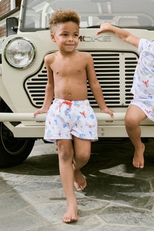 A young boy smiles barefoot in front of a cream-colored vehicle, wearing Roller Rabbit Boys Catch of the Day Shortie Swim Trunk with colorful prints, while another child sits partially visible beside him.