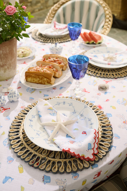 The Catch of the Day Napkin Set Of 4 by Roller Rabbit features white linen napkins with red scalloped edges, showcasing playful sea, beach, and ice cream prints in blue, red, and yellow. The napkins are arranged slightly overlapping.