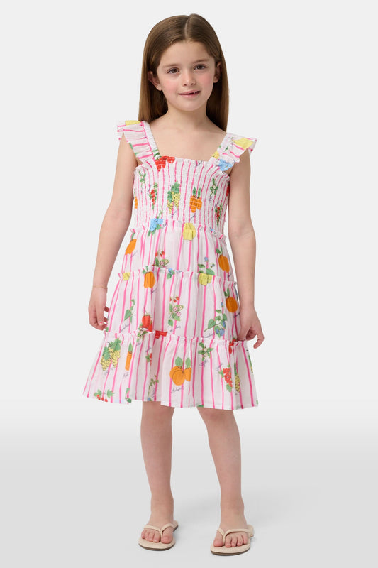 A young girl with straight brown hair wears the Roller Rabbit Kids Fruits D'Ete Ceri Dress, a pink and white smocked sundress with ruffled sleeves, and white sandals, standing and smiling against a light gray background.