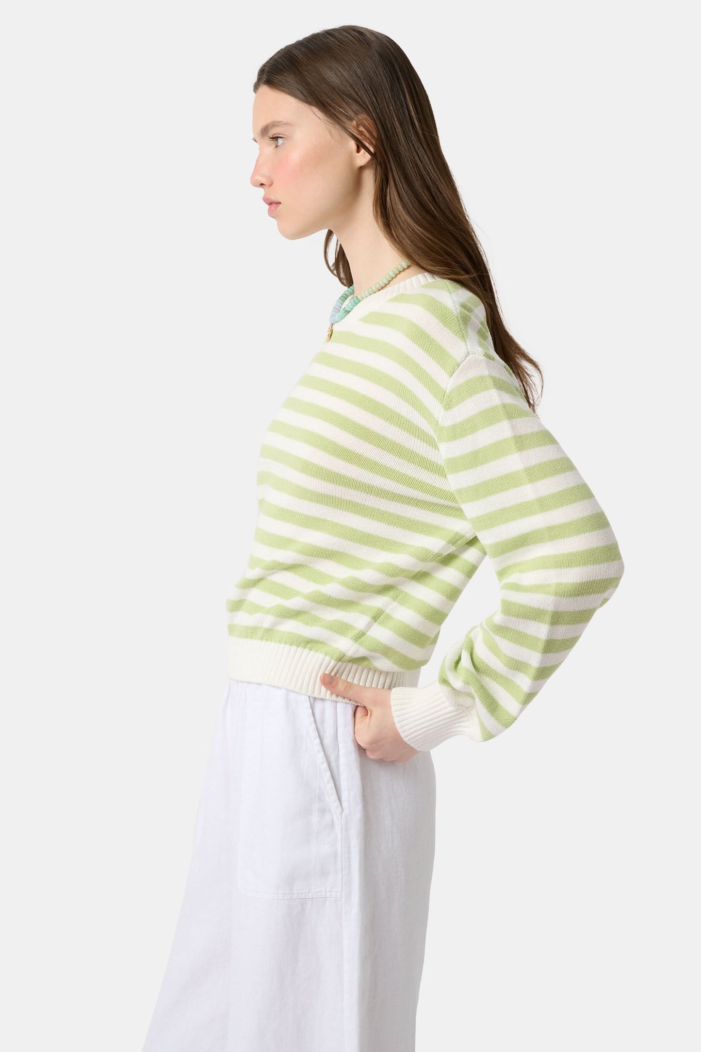 A woman with straight brown hair wears the Roller Rabbit Striped Hadley Sweater in sage green and white stripes, paired with white high-waisted pants and a beaded necklace, standing against a plain background and looking at the camera.