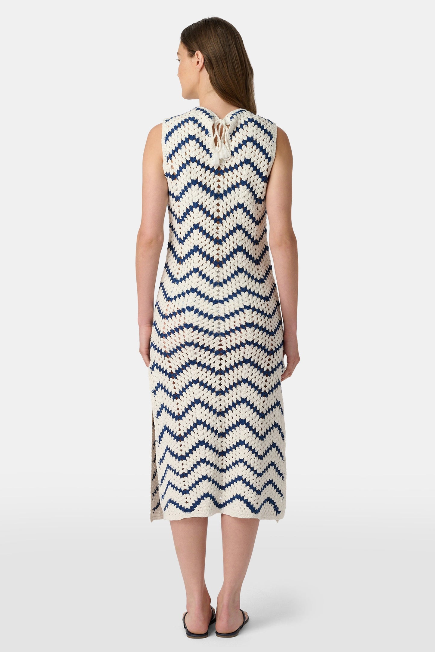 A woman wears the Roller Rabbit Zig Zag Irene Cover Up, a sleeveless, ankle-length crochet maxi dress with a blue and white zig zag pattern. She pairs it with black sandals and chunky striped bracelets, her straight brown hair worn loose.
