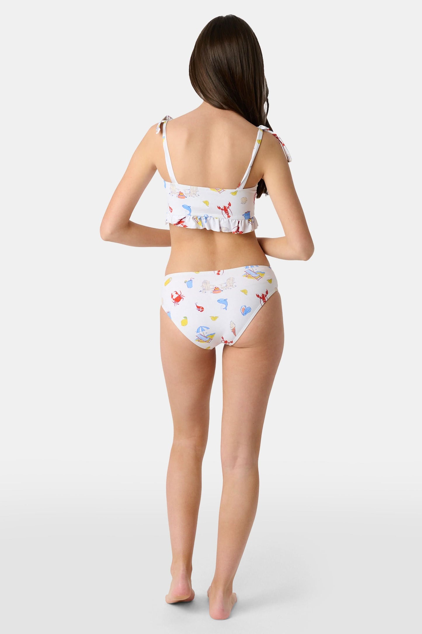 A girl wears the Roller Rabbit Kids Ruffle Catch of the Day Cora Bikini, a white two-piece with a playful print, ruffle trim top, and tie straps, standing against a plain light background.