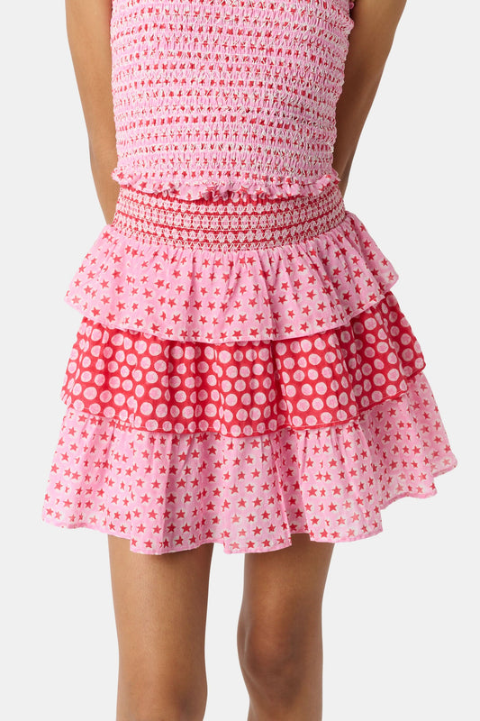 A child wears the Roller Rabbit Kids Starry Dot Sami Skirt in pink and red with star and polka dot patterns, paired with a pink smocked top, standing against a plain light background.
