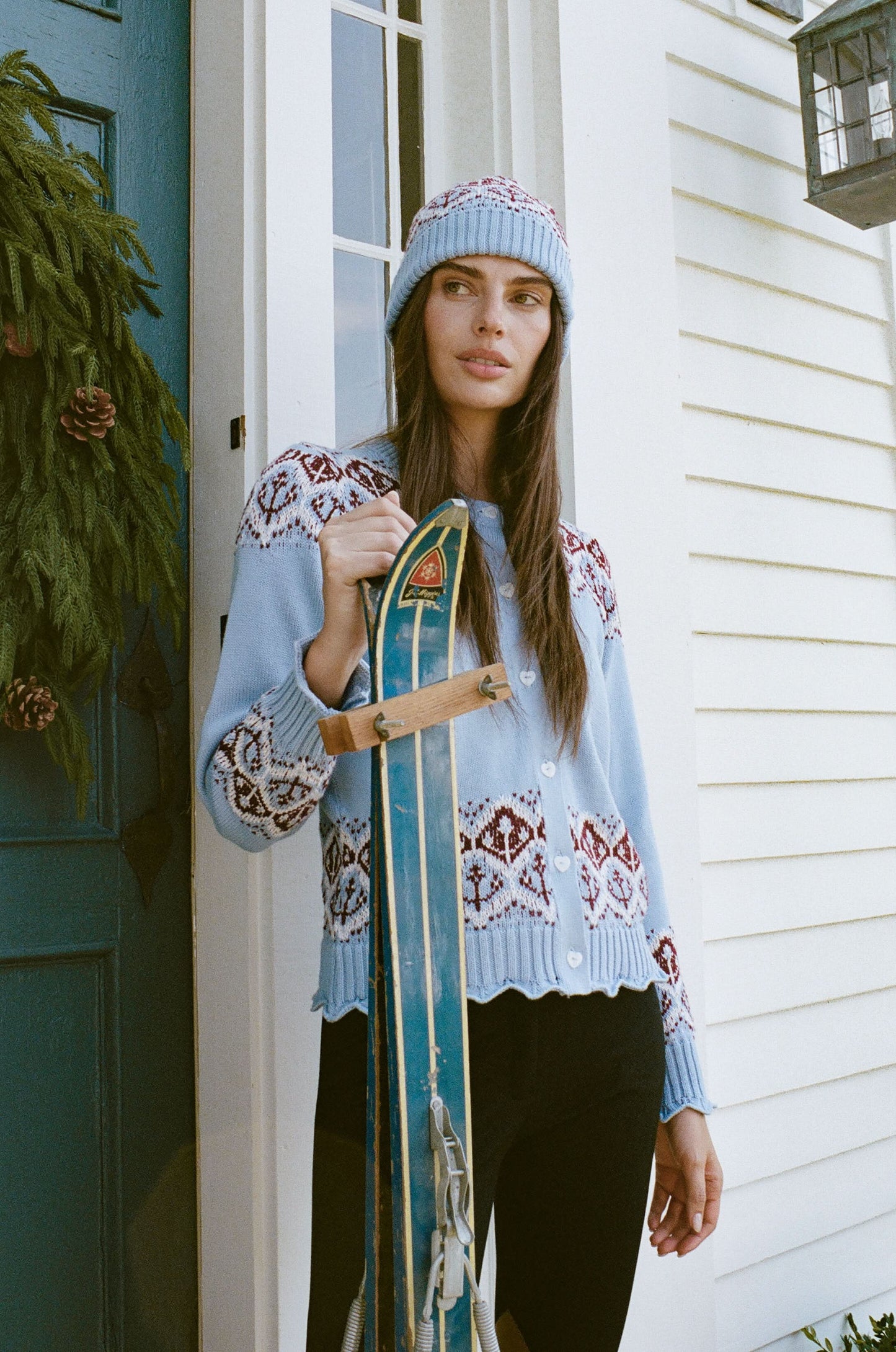 A young woman with fair skin and light makeup gazes at the camera, one hand on her hat, wearing a light blue Roller Rabbit Jemina Fairisle Beanie with a red and white pattern that matches her sweater.