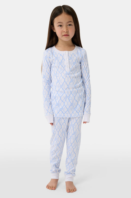 A young girl with long brown hair stands barefoot in Roller Rabbit Kids Seaside Knots Juniper Pajamas, featuring matching white and light blue knot print on soft Pima fabric with long sleeves and button details, against a plain light background.