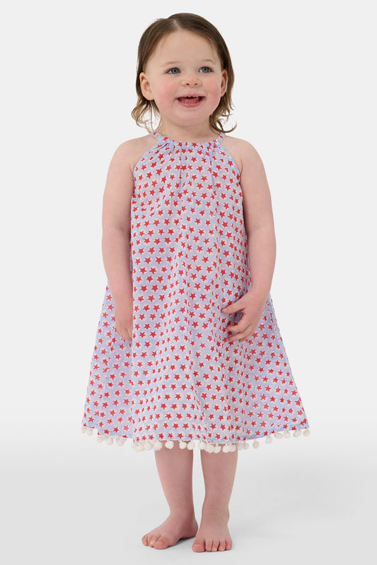 A young girl with long brown hair stands facing forward in the Roller Rabbit Kids Starry Dot Carrie Cover Up—a sleeveless, knee-length white dress featuring a red dotted pattern and pom-pom trim. She has a neutral expression and a light background behind her.