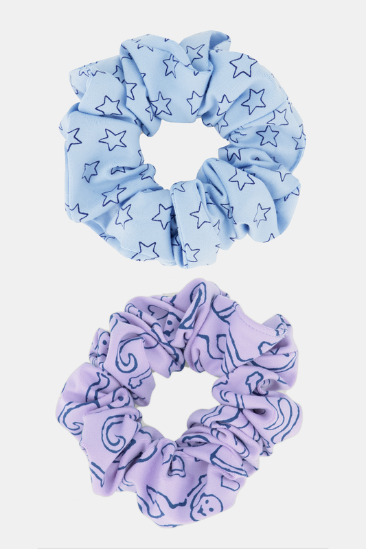 The Assorted Scrunchies Set Of 2 features a light blue scrunchie with dark blue stars and a lavender one with dark blue moons and abstract shapes—fun, stylish hair accessories for any look.