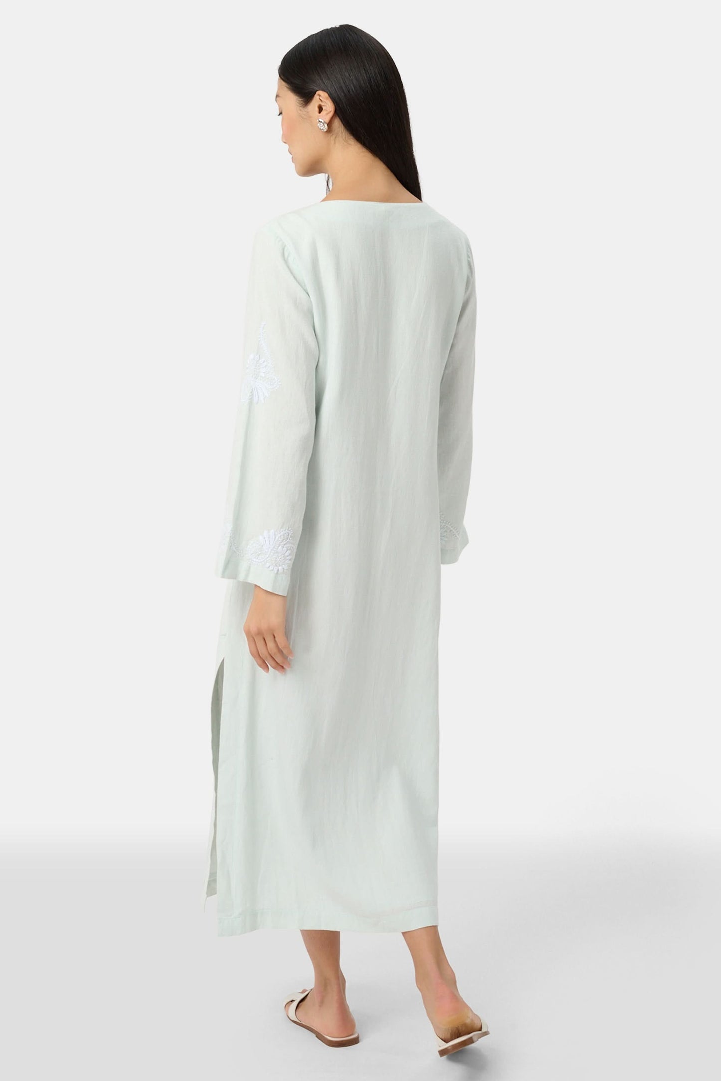 A woman faces forward wearing the Roller Rabbit Vanna Embroidery Rachel Caftan—a long, light green linen dress with subtle white embroidery and long sleeves—paired with white sandals against a plain light gray background.