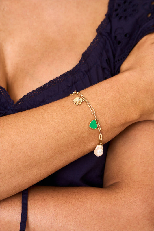 A person in a dark blue, eyelet-patterned top crosses their arms, showcasing the Roller Rabbit Celine Heart Bracelet adorned with candy-colored charms like a green heart, flower, and pearl.