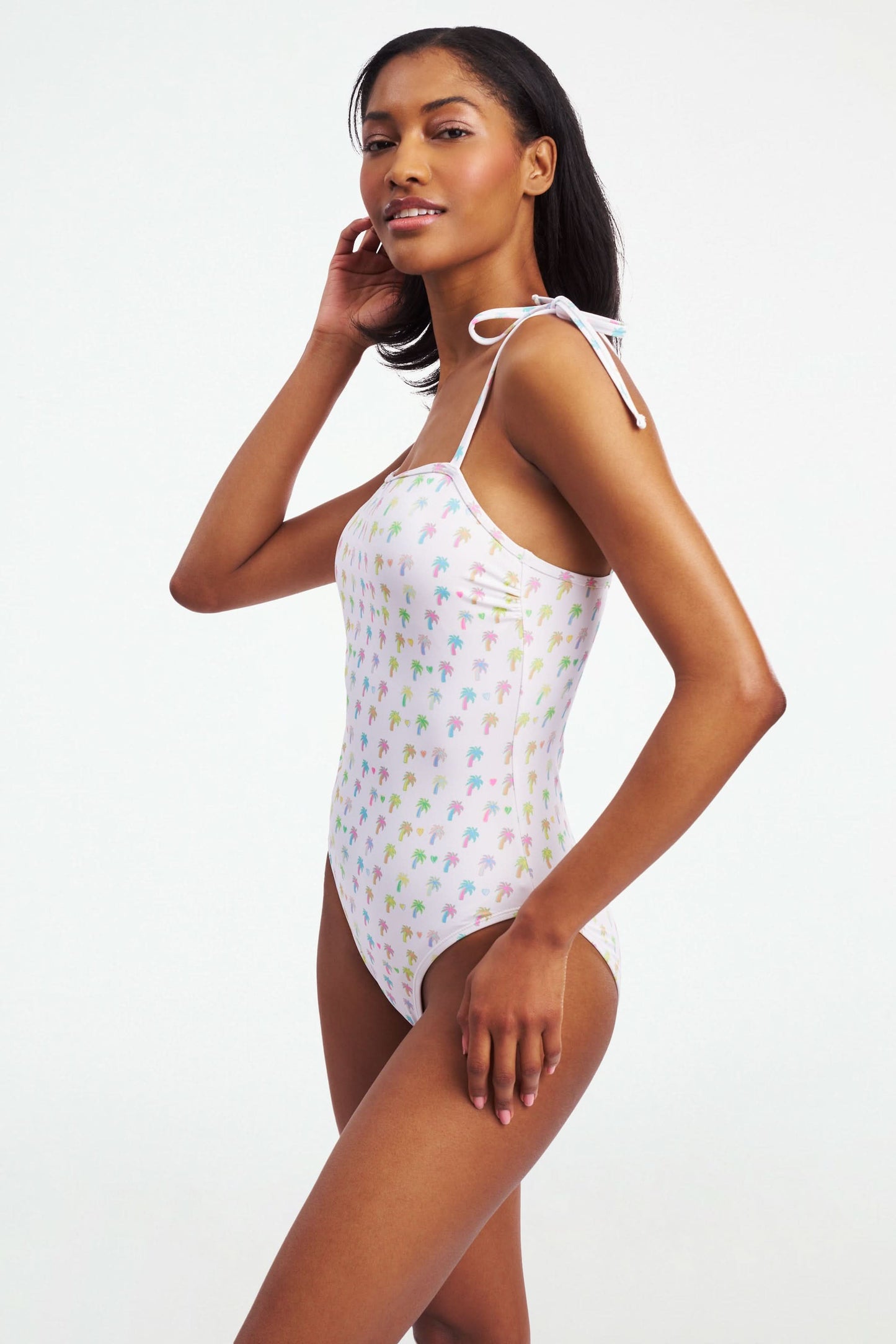 The Roller Rabbit Palm Dreams Cove One Piece is a white swimsuit with vibrant palm tree patterns and adjustable tie shoulders in matching fabric.
