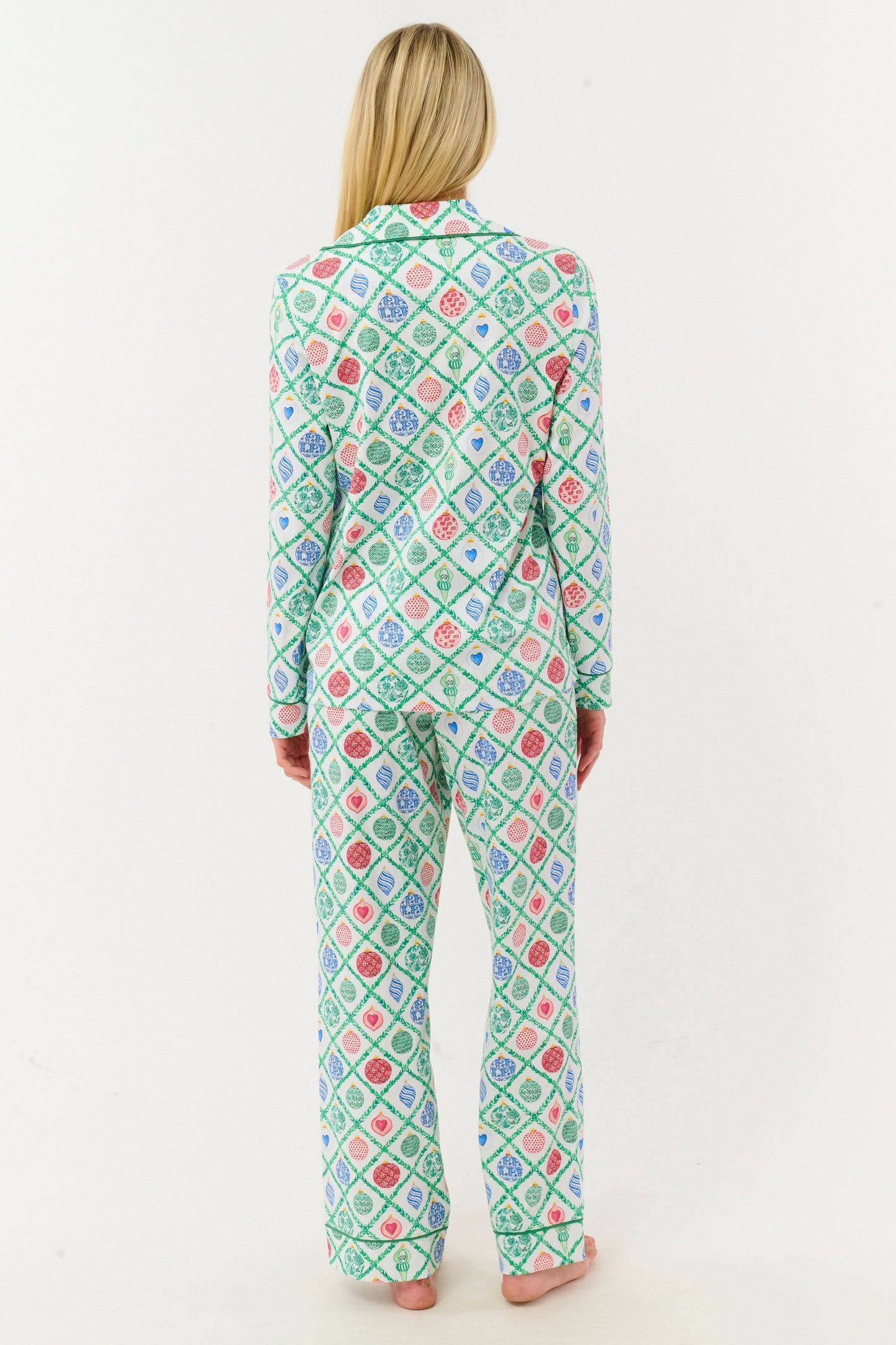 A woman stands barefoot against a white background, wearing Roller Rabbit Ornament Trellis Long Sleeve Polo Pajamas—holiday PJs with a blue, green, and pink geometric diamond print, made from 100 Pima cotton.