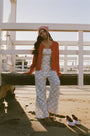 Someone stands against a plain white background wearing Roller Rabbit's Raspberry Garden Monroe Pant—loose white loungewear pants with a pink and green floral print—paired with maroon slippers adorned with white floral details.