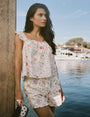 A woman wears the Roller Rabbit Baroque Floral Saylor Set—light cotton loungewear with a sleeveless ruffled top and loose shorts—standing with one hand in her pocket against a plain background.