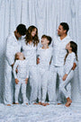 A man stands barefoot on a white background wearing Roller Rabbit's Mens Starry Nights Lattice Long Sleeve Polo Pajamas, featuring light blue and white patterns. He smiles with one hand in his pocket, highlighting the comfort of these matching PJs.
