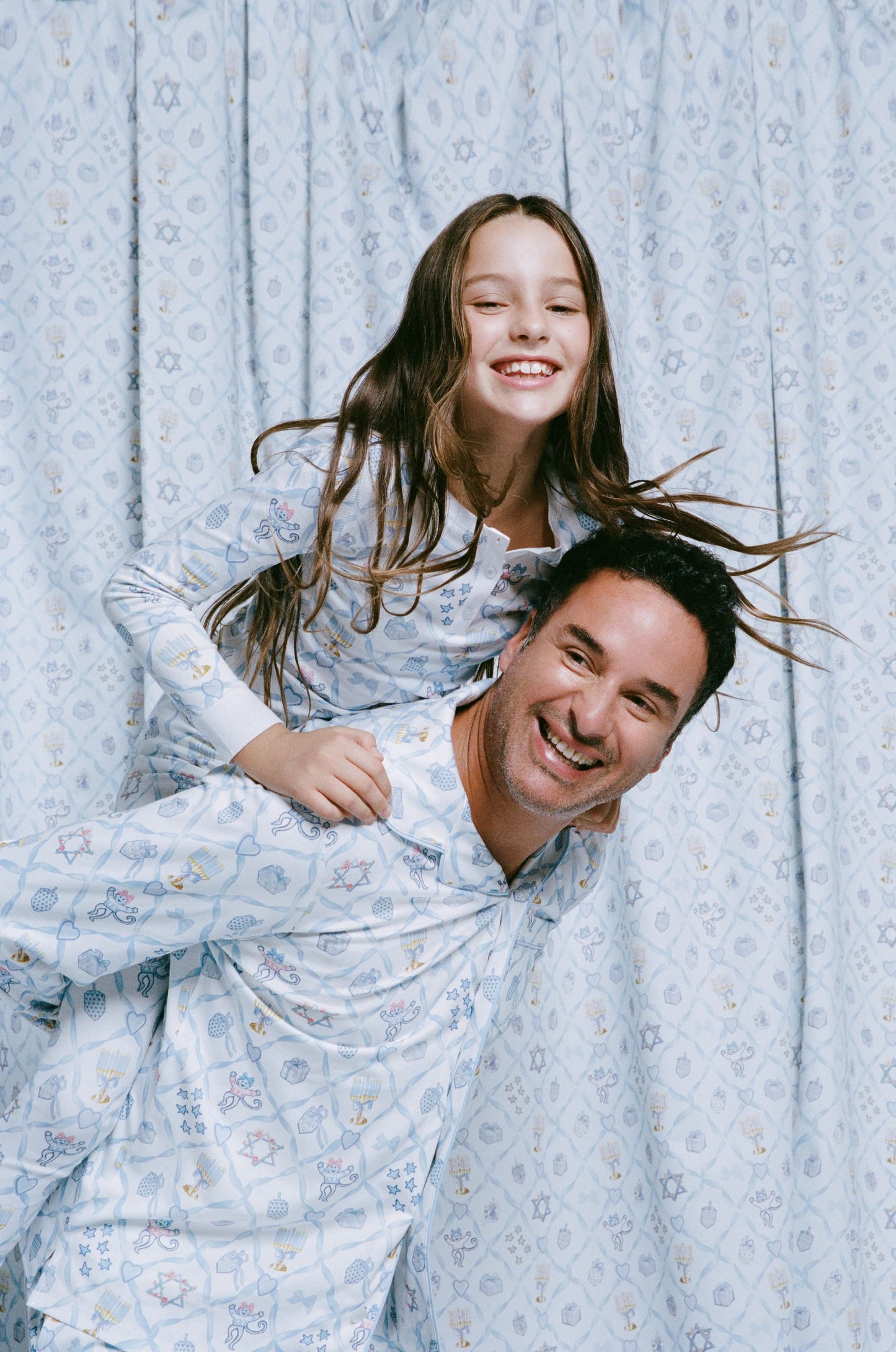 A man stands barefoot on a white background wearing Roller Rabbit's Mens Starry Nights Lattice Long Sleeve Polo Pajamas, featuring light blue and white patterns. He smiles with one hand in his pocket, highlighting the comfort of these matching PJs.