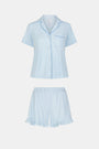 A woman wears Roller Rabbit's Hearts Tonal Wyatt Polo Pajamas—light blue, short-sleeved Pima cotton with a collar and chest pocket—paired with matching ruffled shorts, standing against a plain light background.