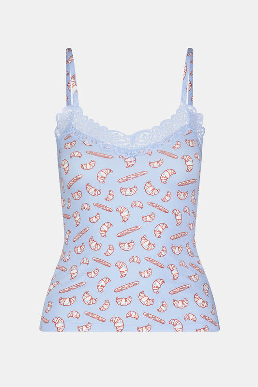 A woman models the Roller Rabbit Breakfast In Bread Perry Cami, featuring scalloped lace trim and a playful croissant print. The photo is cropped from shoulders to hips against a plain white background.