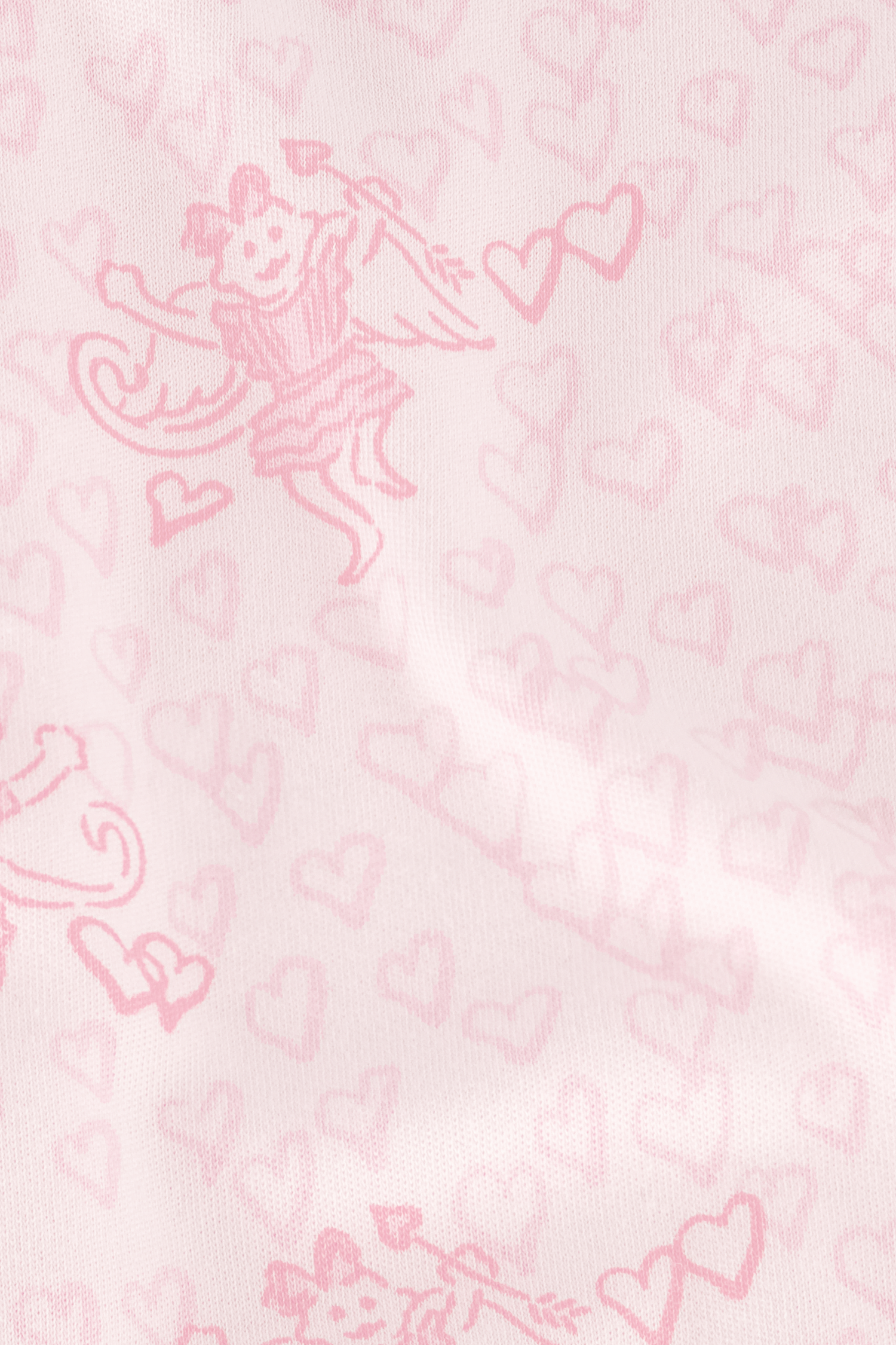 A young woman wearing the Roller Rabbit Cupid's Kiss Boden Pant and fluffy slippers stands in front of a pastel pink, candy-themed counter adorned with jars of sweets and heart decorations.