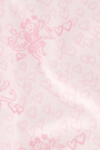 A young woman wearing the Roller Rabbit Cupid's Kiss Boden Pant and fluffy slippers stands in front of a pastel pink, candy-themed counter adorned with jars of sweets and heart decorations.