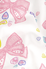 A woman stands smiling against a plain light background, wearing Roller Rabbit’s Doily Beaux Polo Pajamas—a short-sleeved, collared white set with pink bow patterns and small colorful shapes.