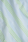 A woman with long brown hair wears the Roller Rabbit Positano Stripe Cooper Set, featuring a loose, sleeveless cotton poplin top with ruffle trim and drawstring, and elastic-waist shorts in white fabric with light green vertical stripes.