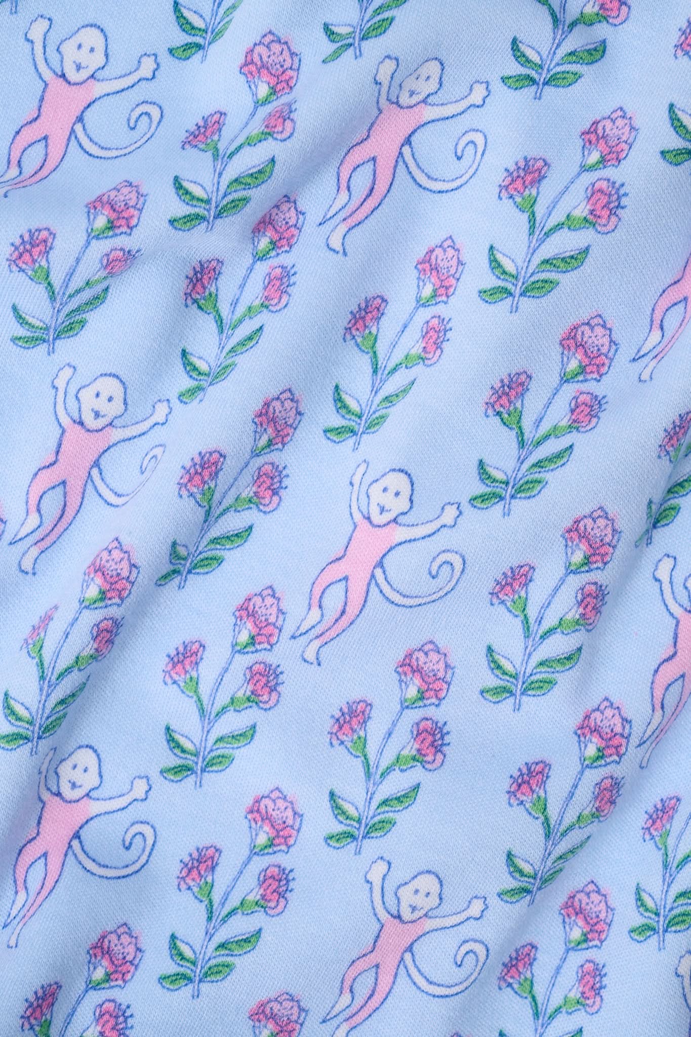 A woman with long curly hair wears the Rosa Ditsy Monkey Alexa Set by Roller Rabbit, a light blue Pima cotton pajama set featuring a ditsy monkey print. The set includes a henley-style top and matching shorts. She stands against a plain background.