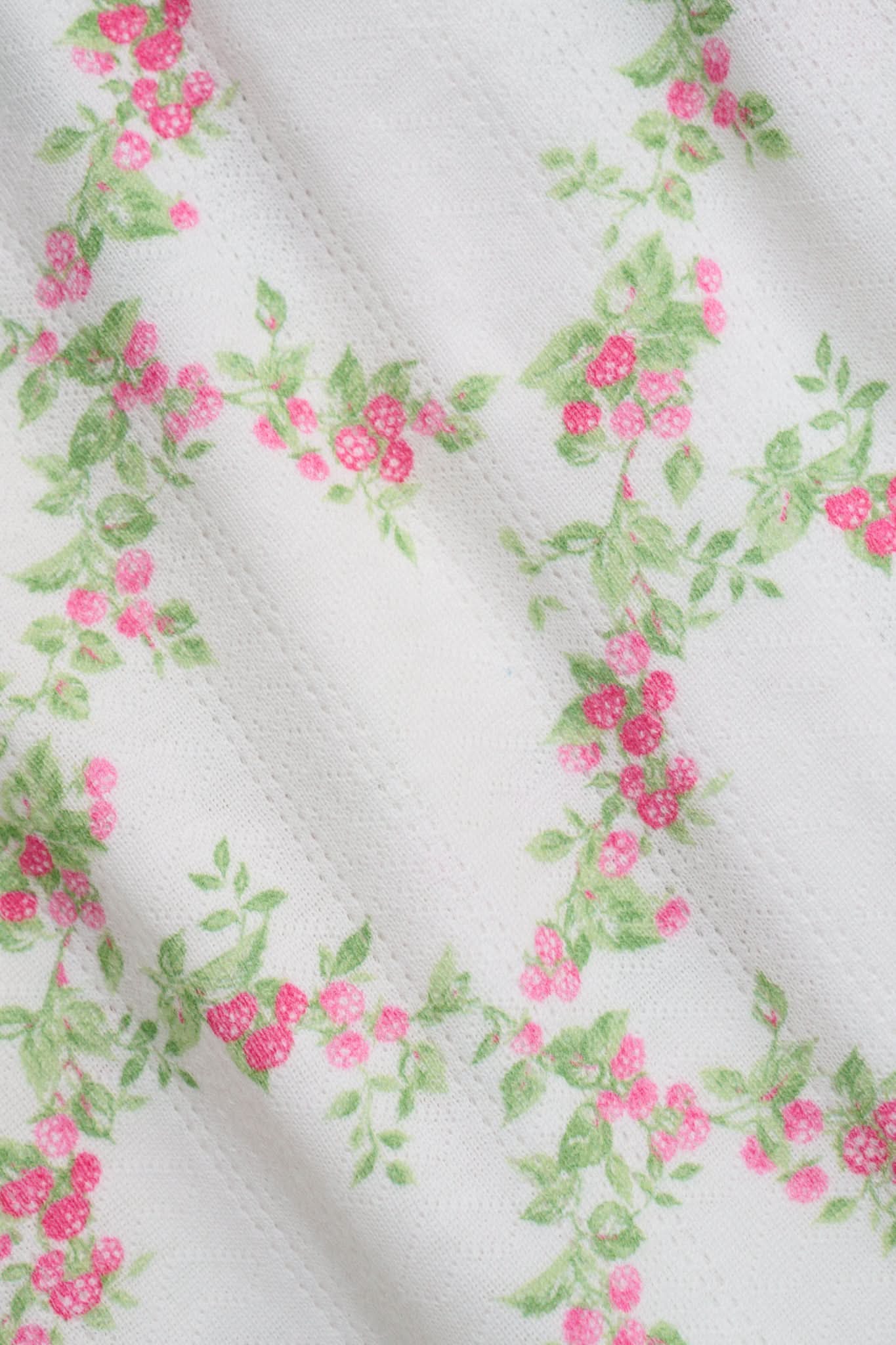 Someone stands against a plain white background wearing Roller Rabbit's Raspberry Garden Monroe Pant—loose white loungewear pants with a pink and green floral print—paired with maroon slippers adorned with white floral details.