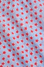 A young girl with long brown hair stands facing forward in the Roller Rabbit Kids Starry Dot Carrie Cover Up—a sleeveless, knee-length white dress featuring a red dotted pattern and pom-pom trim. She has a neutral expression and a light background behind her.