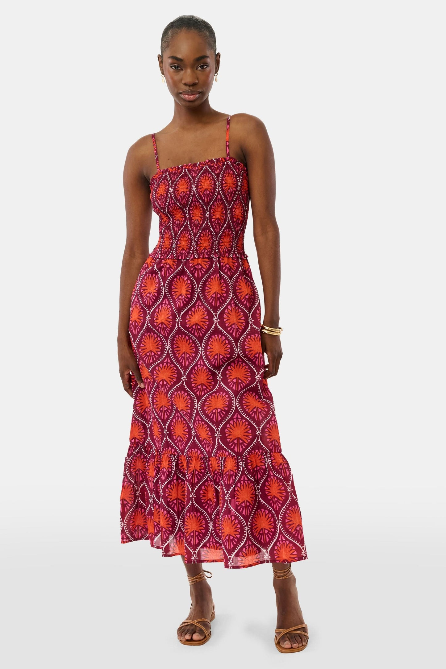 A woman models the Roller Rabbit Palm Fronds Briar Dress, a sleeveless cotton maxi with red, orange, and purple geometric patterns, paired with brown sandals and gold bracelets against a white background.