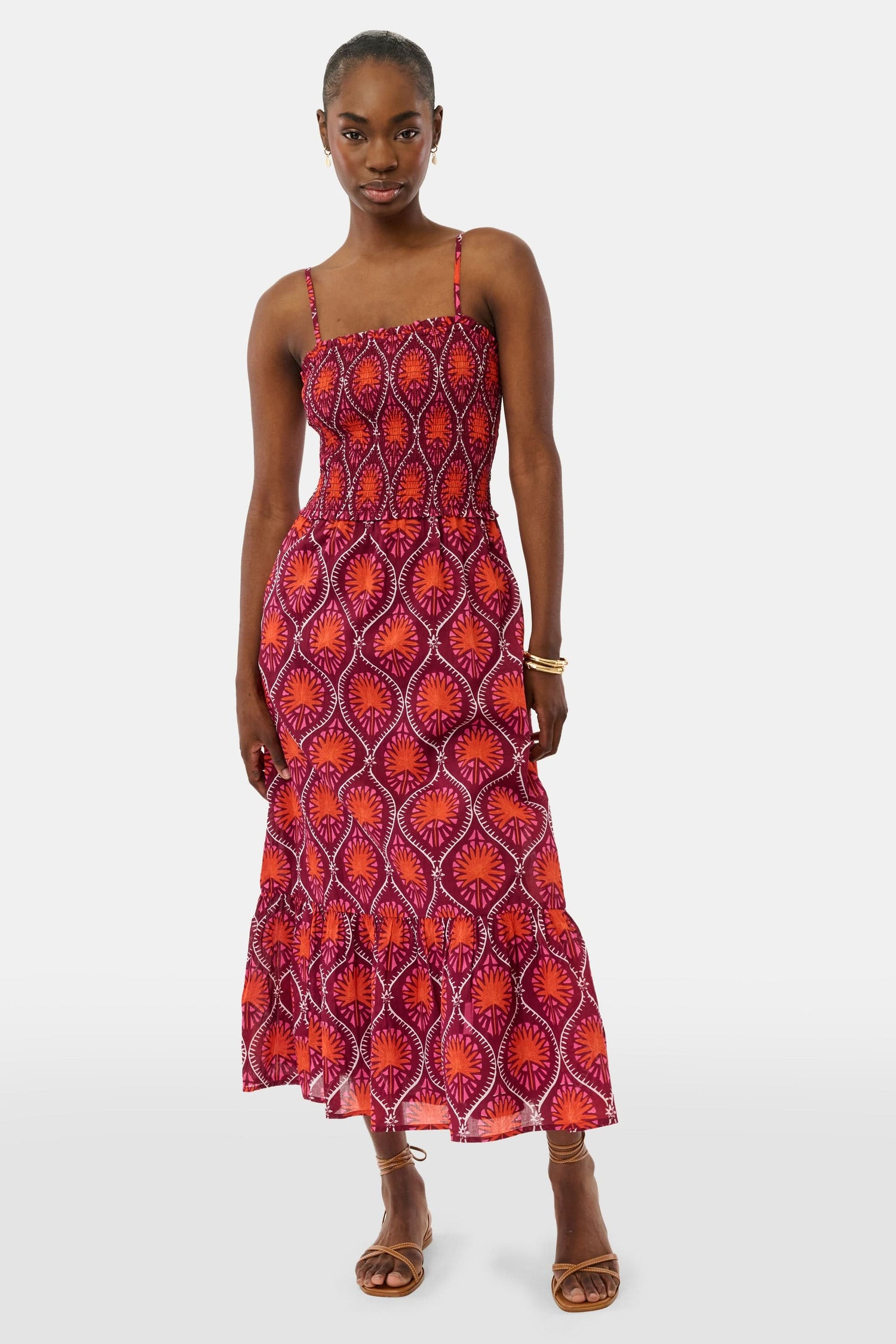 A woman models the Roller Rabbit Palm Fronds Briar Dress, a sleeveless cotton maxi with red, orange, and purple geometric patterns, paired with brown sandals and gold bracelets against a white background.