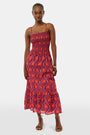 A woman models the Roller Rabbit Palm Fronds Briar Dress, a sleeveless cotton maxi with red, orange, and purple geometric patterns, paired with brown sandals and gold bracelets against a white background.