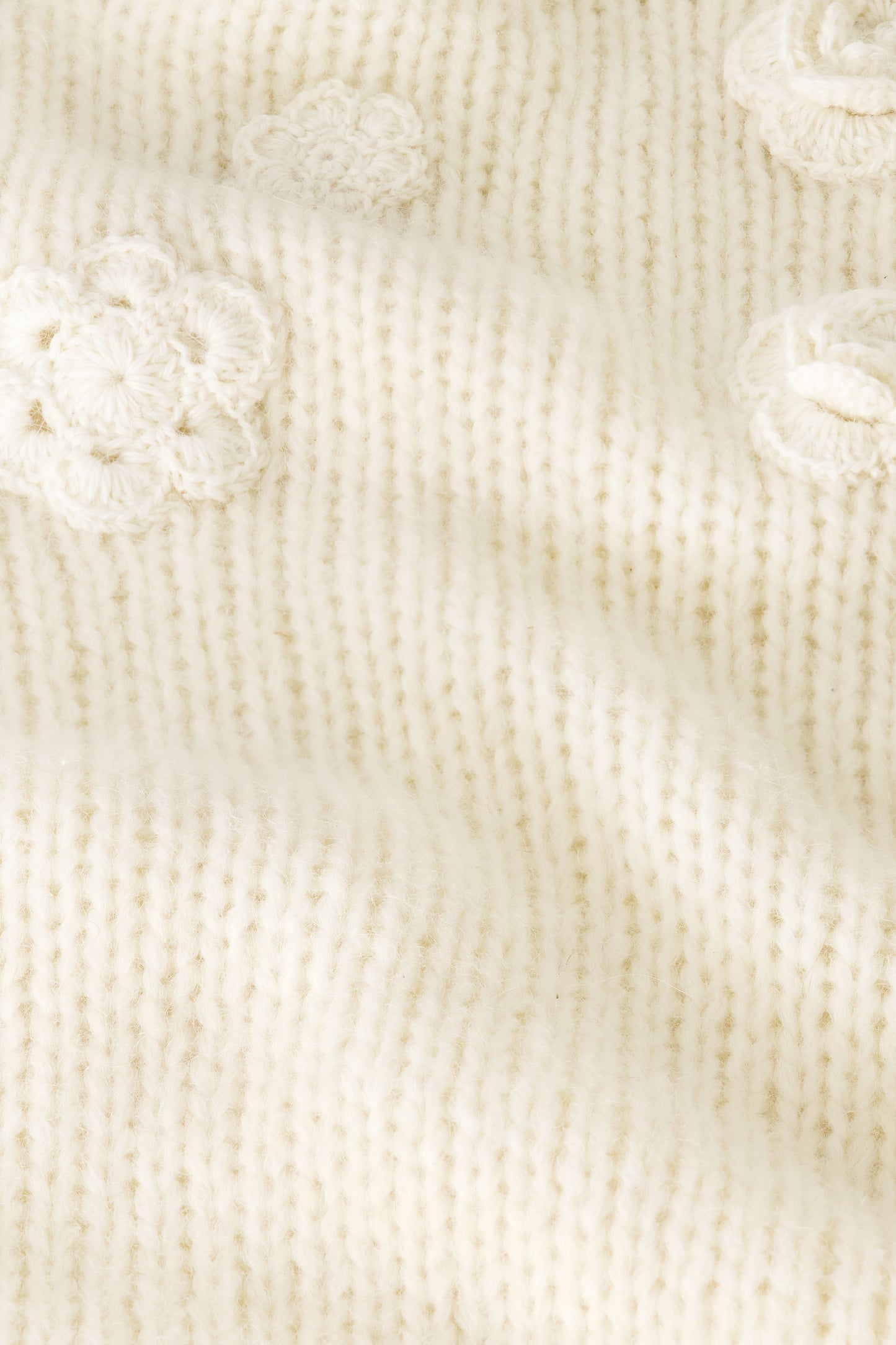 A close-up of the Crochet Floral Quinn Sweater’s soft, cream knit showcases raised crochet floral details and textured stitches, giving it a cozy, delicate look.