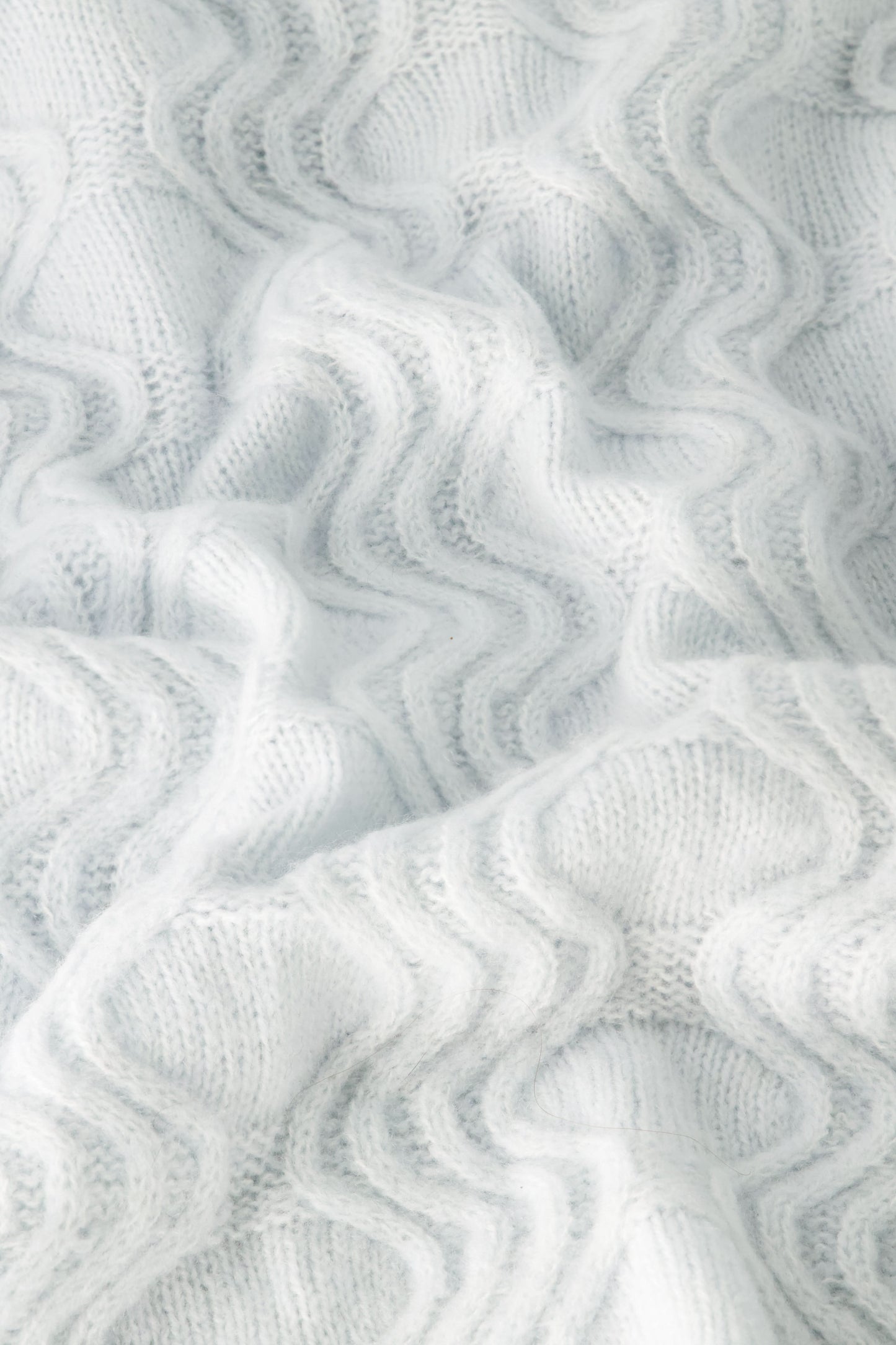 The Wave Knit Ava Sweater features soft, white knitted fabric with a wavy, textured pattern that creates gentle folds and shadows across its surface.