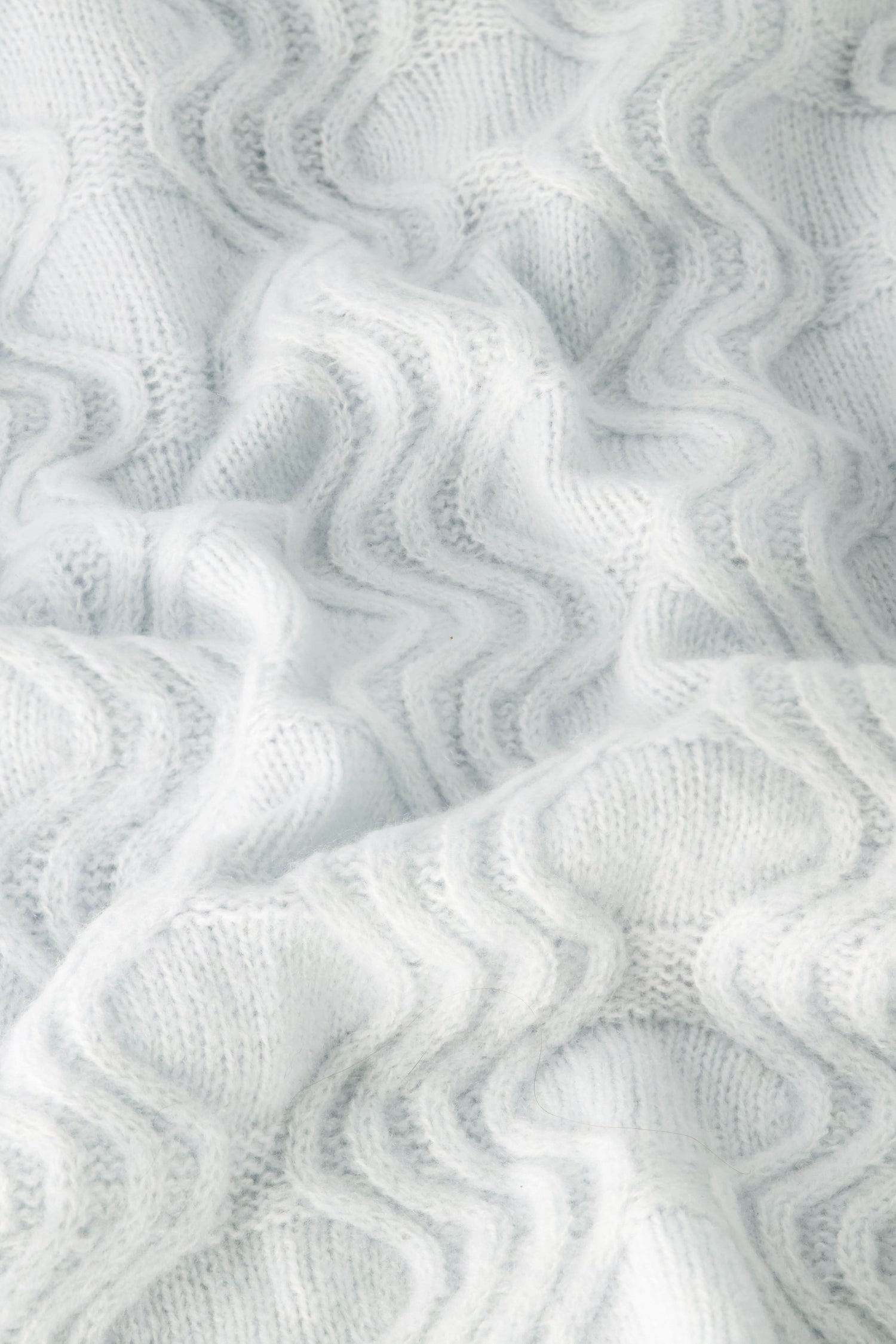 The Wave Knit Ava Sweater features soft, white knitted fabric with a wavy, textured pattern that creates gentle folds and shadows across its surface.