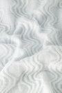 The Wave Knit Ava Sweater features soft, white knitted fabric with a wavy, textured pattern that creates gentle folds and shadows across its surface.