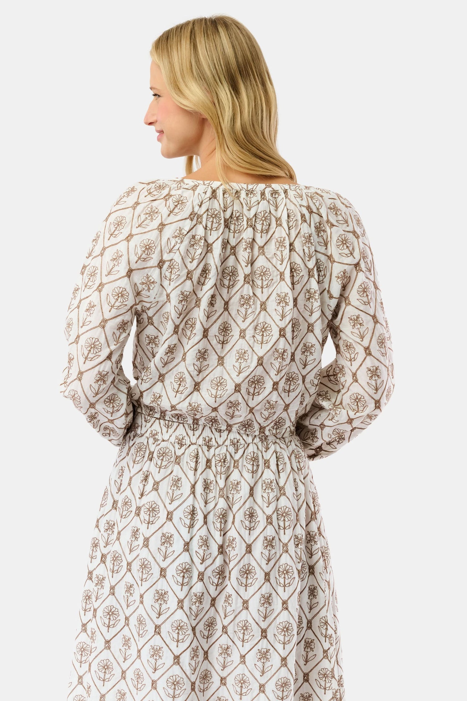 A woman with blonde hair wears the Infinity Floral Embroidery Stevie Top by Roller Rabbit, featuring vintage floral embroidery and a drawstring neckline, standing against a plain white background and smiling softly.